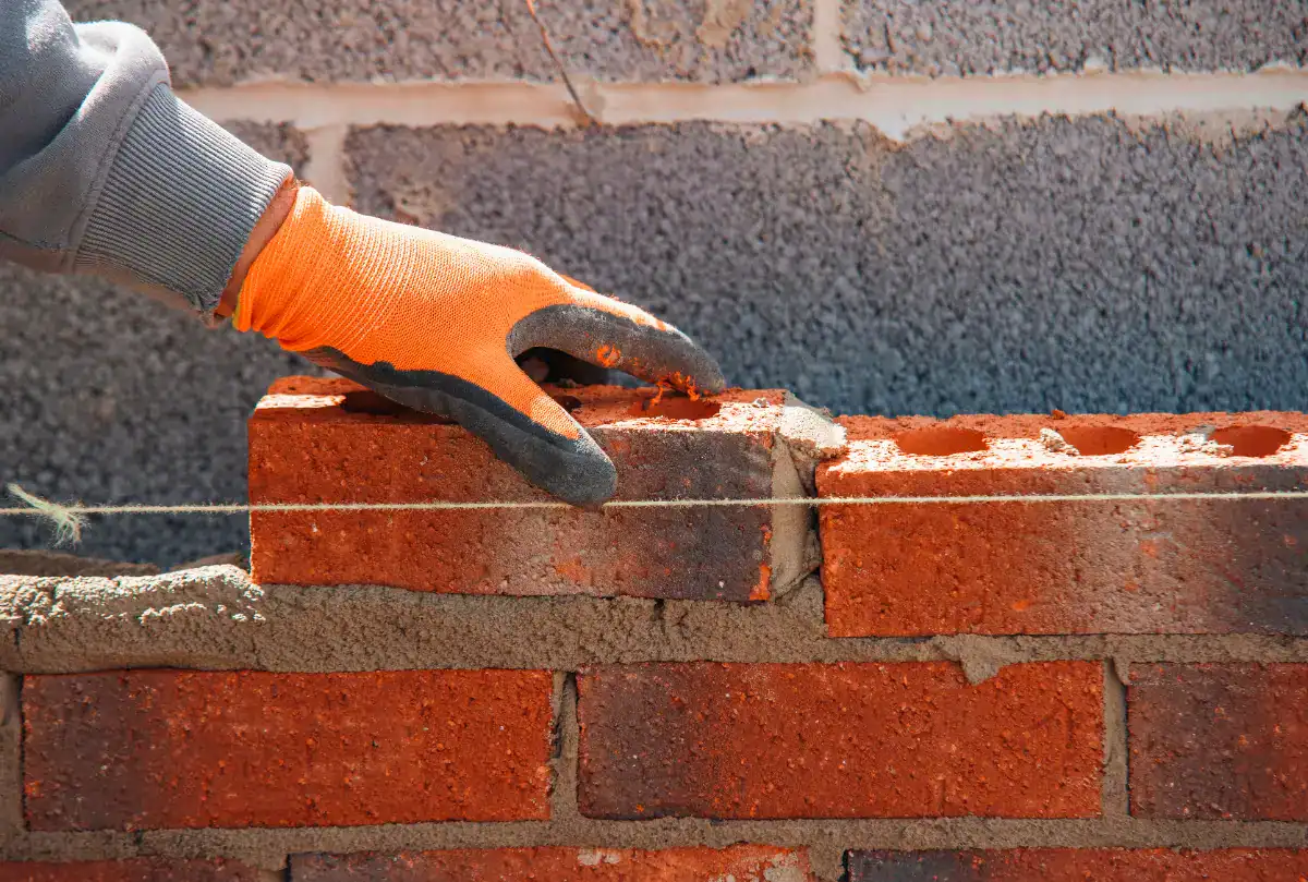 A person wearing an orange work glove places a red brick onto wet mortar in a brick wall, using a taut string for alignment—showcasing the skill of a masonry contractor Nassau County.