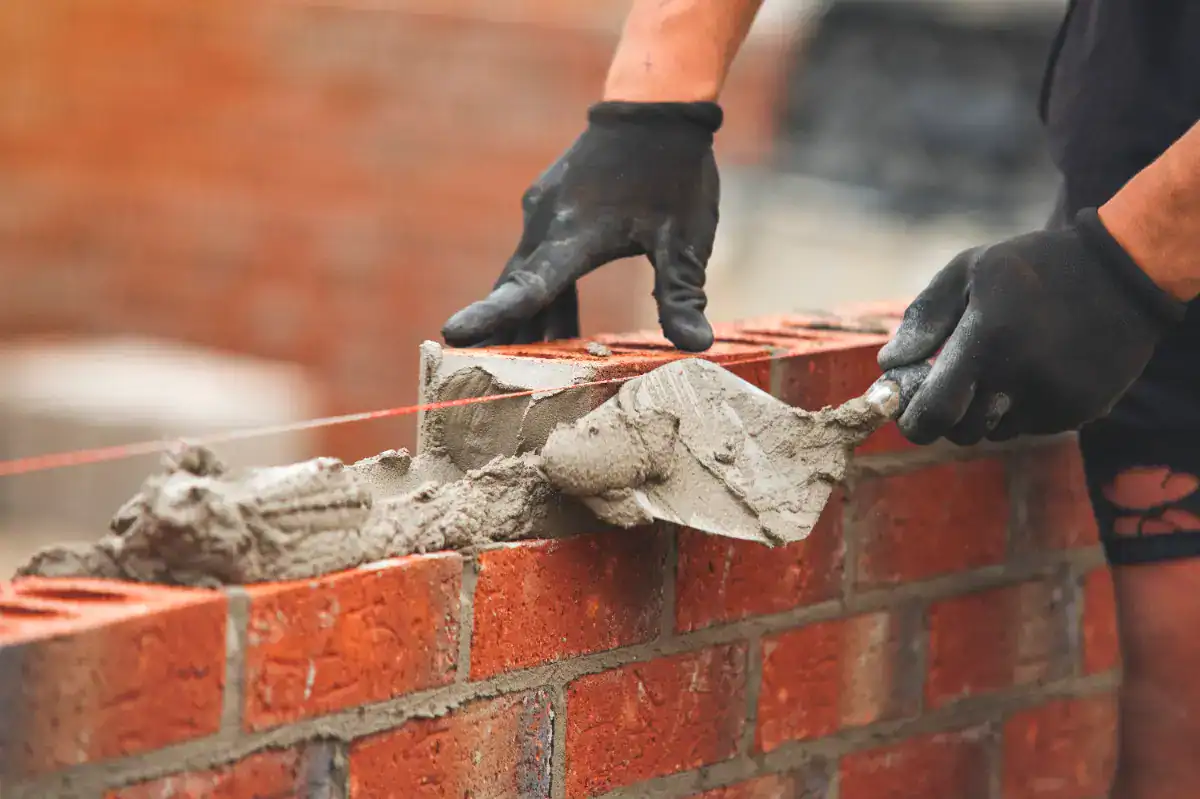 A person wearing black gloves uses a trowel to spread mortar on red bricks while constructing a brick wall. A string line ensures the wall is straight—a common scene for an expert masonry contractor Nassau County, NY.