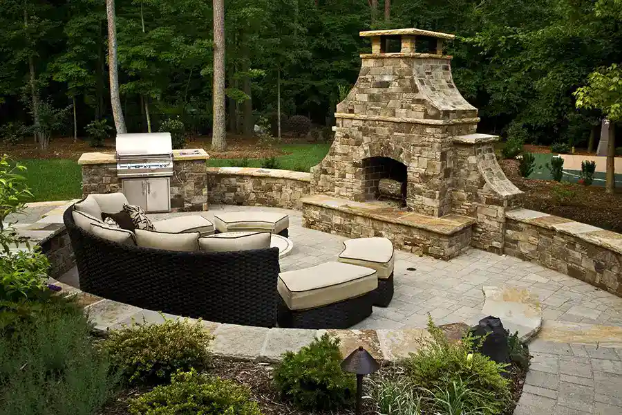 A circular patio with cushioned wicker seating surrounds a fire pit. Designed by a top masonry contractor Nassau County, the space features a stone fireplace and stainless grill, bordered by lush greenery—perfect for relaxing outdoors in NY.