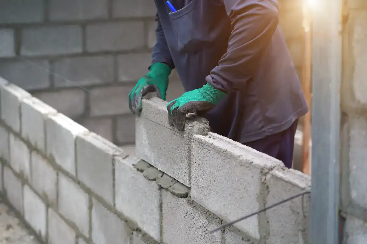 A masonry contractor Nassau County, NY, wearing green gloves and a dark outfit is laying concrete blocks to build a wall, with sunlight shining in the background.
