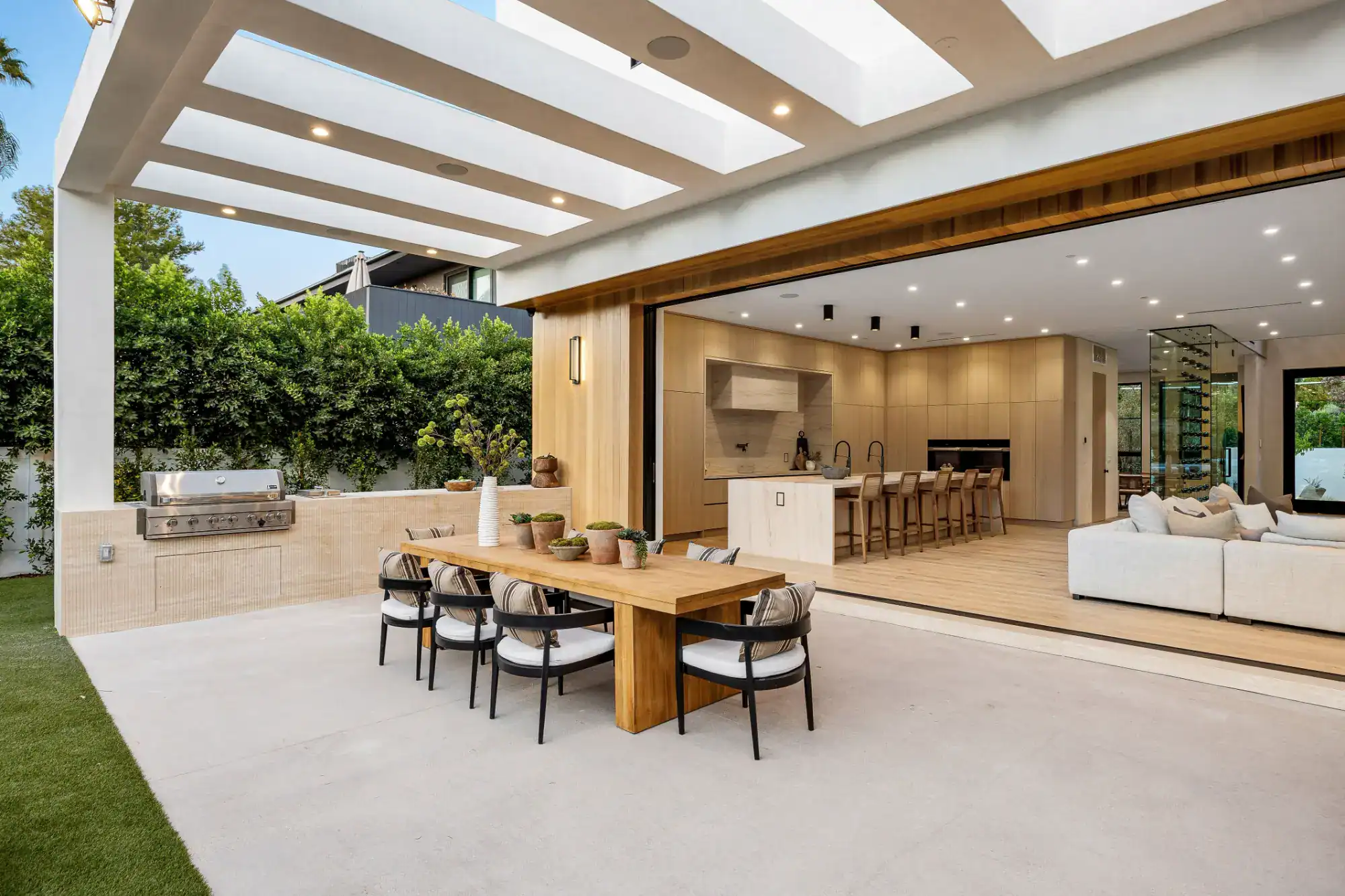 Modern outdoor patio by a leading masonry contractor Nassau County, featuring a wooden dining table, built-in barbecue grill, and open sliding doors leading to a spacious kitchen and living room with light wood finishes and neutral decor.