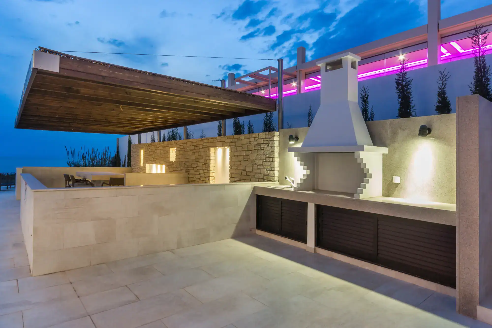 Modern outdoor kitchen with a built-in grill, stone walls crafted by a top masonry contractor Nassau County, and a wood-paneled pergola ceiling. Pink LED lights illuminate the area, while spotlights highlight the cooking space at dusk.