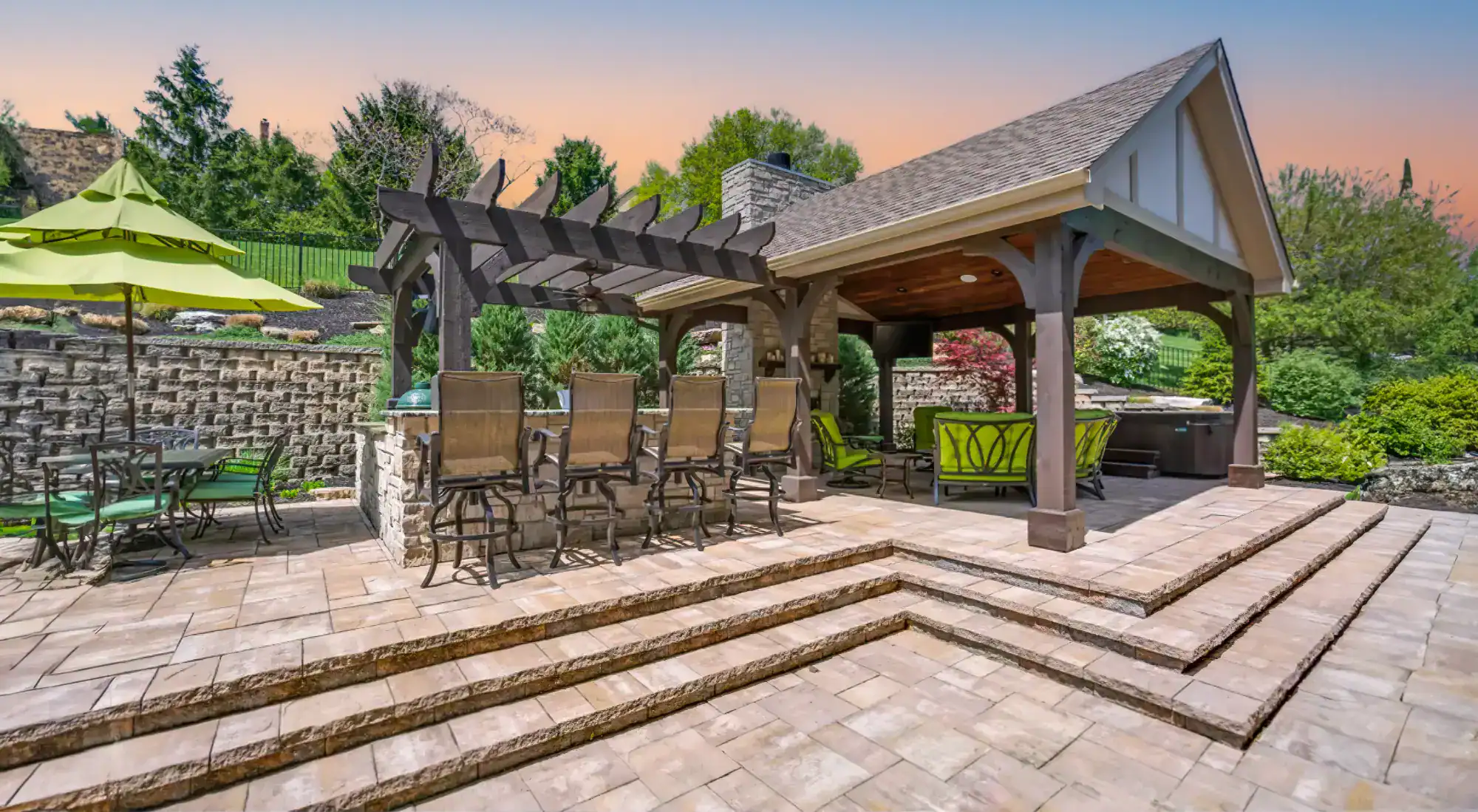 Spacious outdoor patio at sunset with a pergola, high-top bar seating, a covered lounge area, expertly crafted stone steps by a masonry contractor Nassau County, and lush greenery in the background.