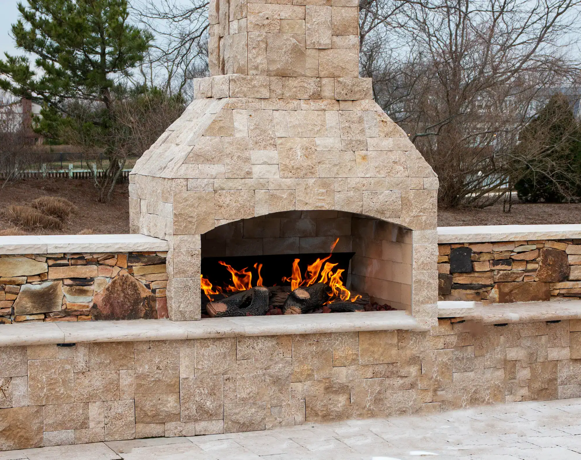 Outdoor stone fireplace with a burning fire, set in a patio area crafted by a skilled masonry contractor Nassau County, surrounded by stone walls, with bare trees and shrubs in the background.