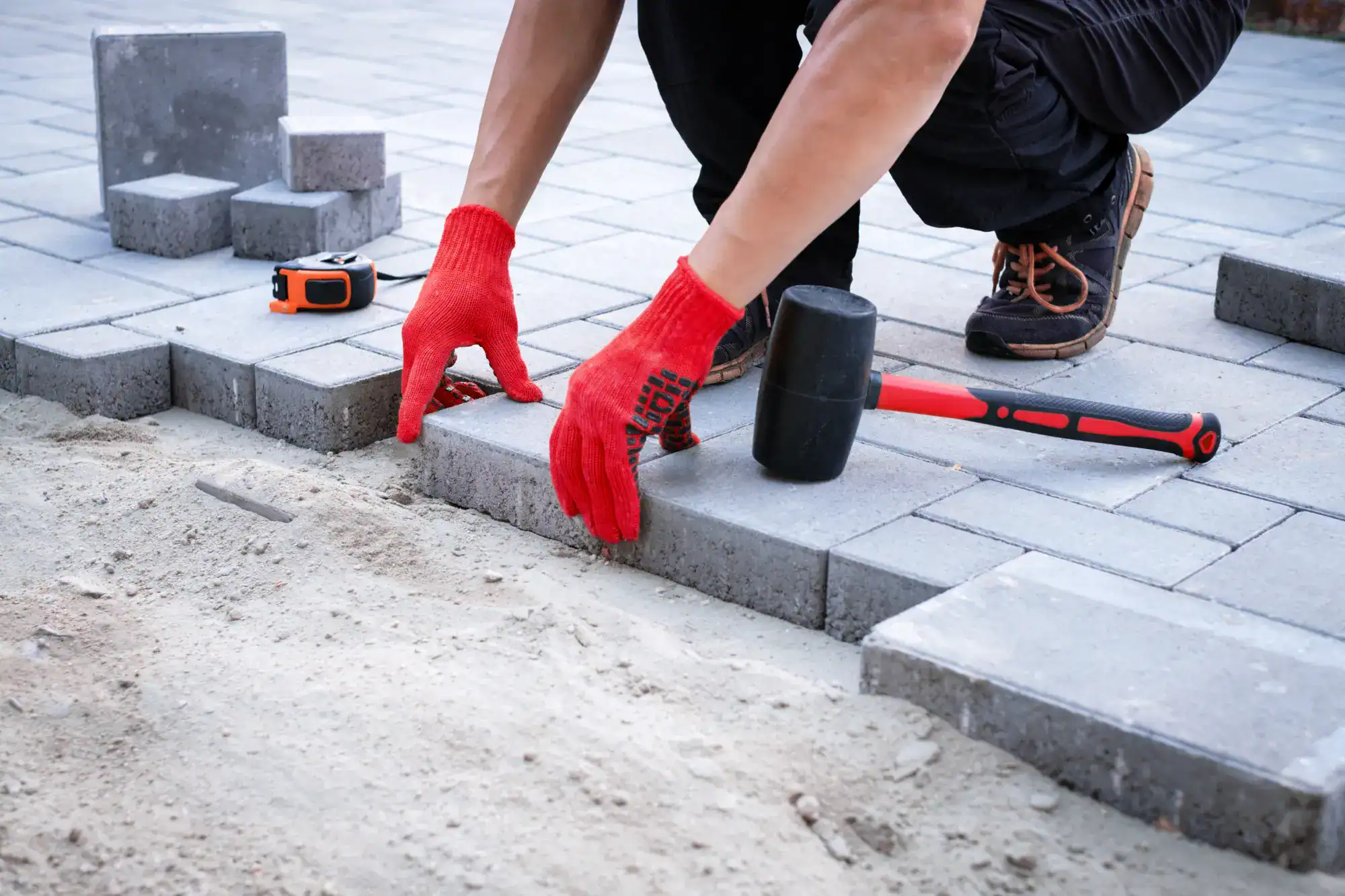 A person in red gloves and black work boots, likely a masonry contractor Nassau County, is laying gray pavers on sand with a rubber mallet and tape measure, working diligently on constructing a paved walkway or patio.