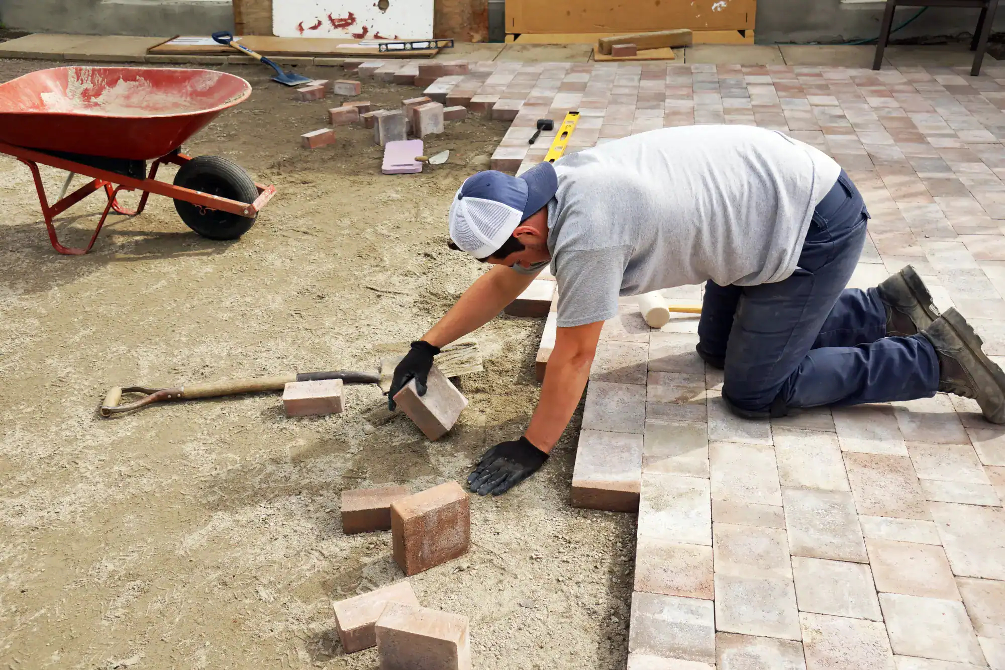 A masonry contractor Nassau County, NY kneels on the ground, wearing gloves and a cap, carefully placing brick pavers for a patio. Nearby, a wheelbarrow, shovel, and spirit level rest on the unfinished surface.