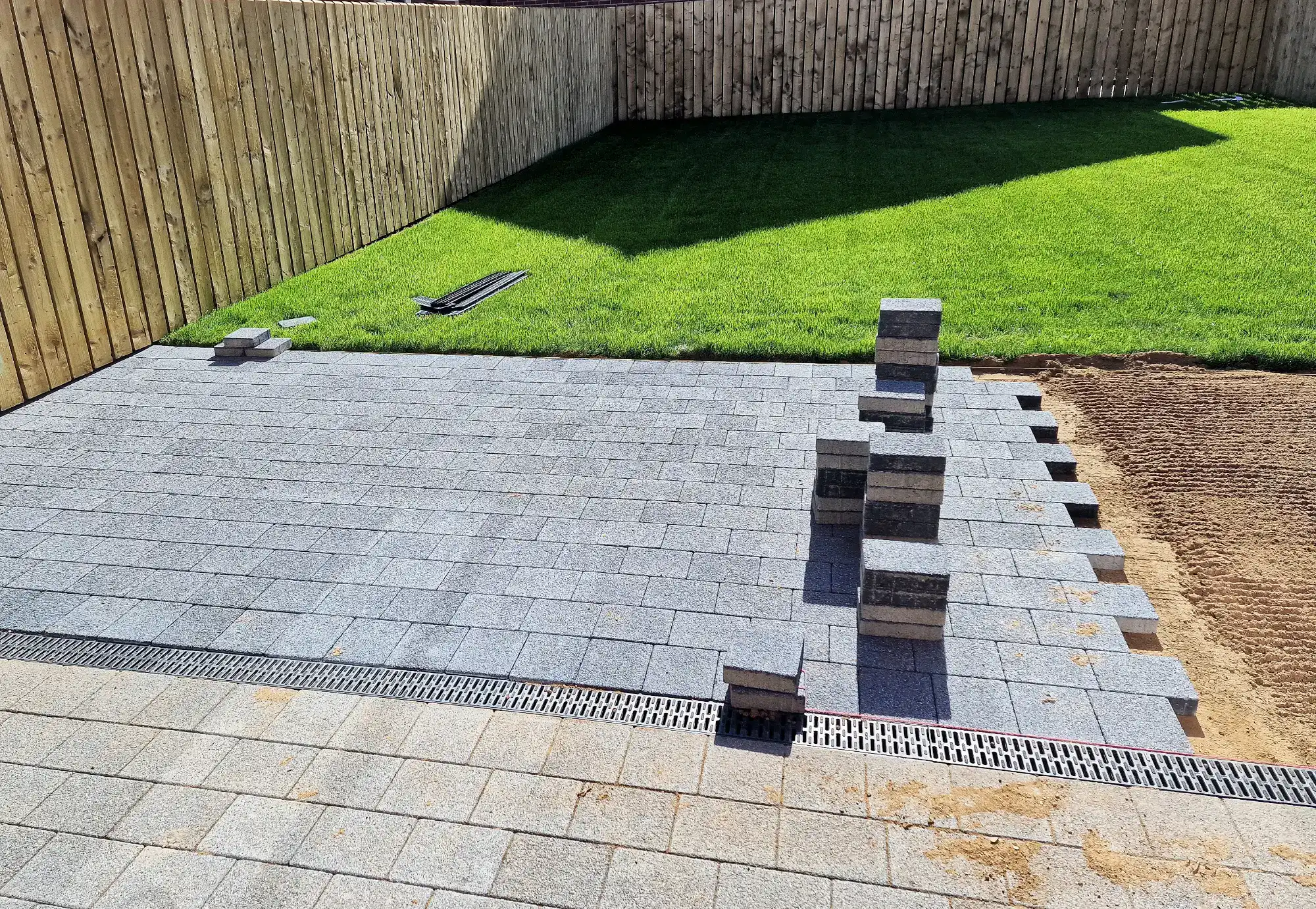 Gray paving stones being laid in a backyard by a masonry contractor Nassau County, NY, with some stacked in piles on the partly finished surface. A section of green lawn and a wooden fence are in the background. Sand and tools are visible on the side.