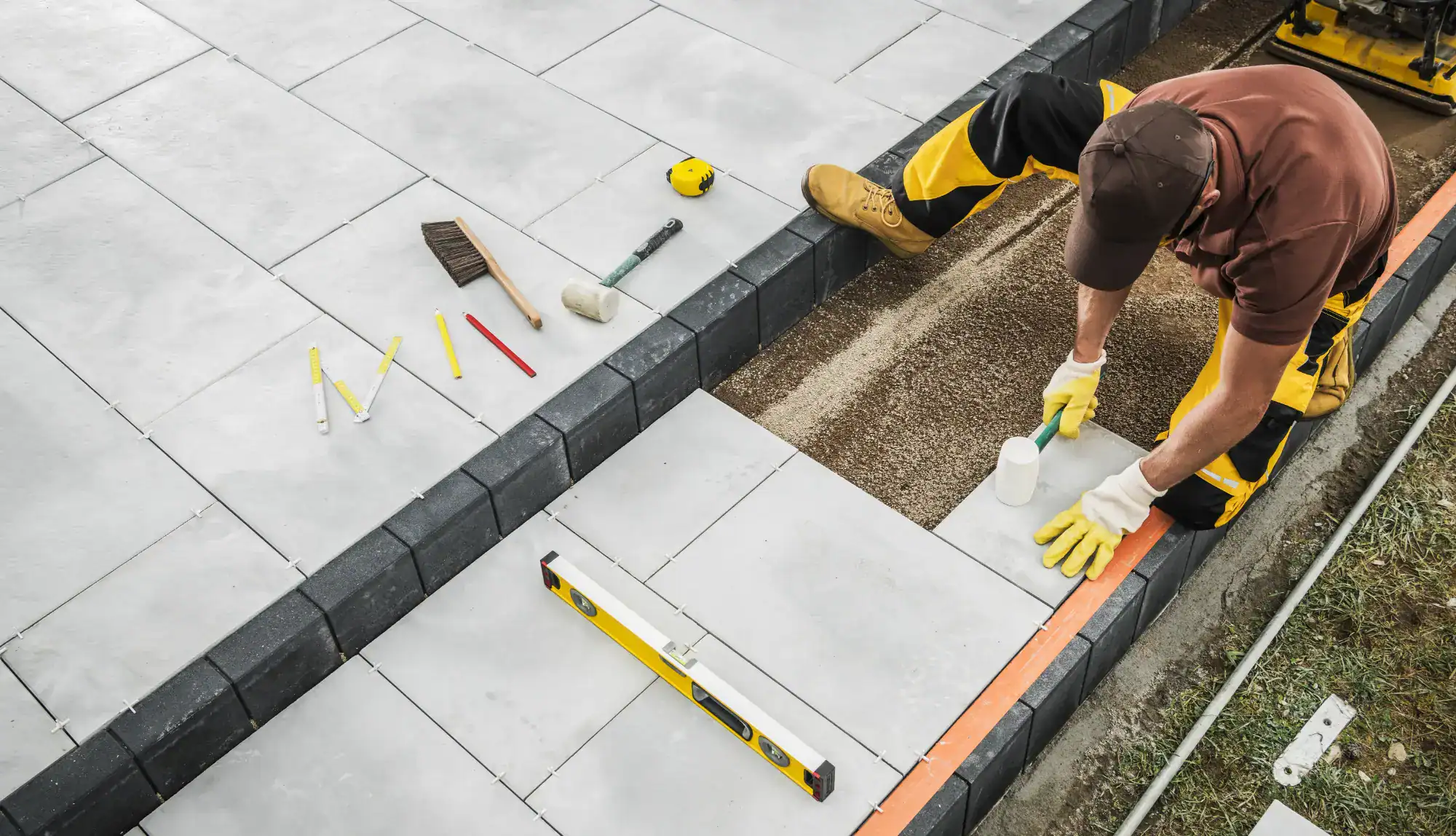A worker wearing gloves and yellow work pants uses a rubber mallet to install concrete paving slabs, with various tools like a level, tape measure, and broom nearby—typical of a skilled masonry contractor Nassau County, NY.