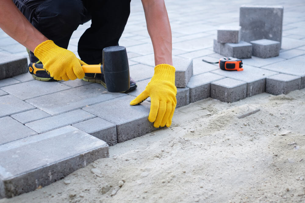 A masonry contractor Nassau County uses yellow gloves and a rubber mallet to lay gray paving stones on sand, creating a patio or walkway. Nearby, a tape measure and extra pavers are ready for use.