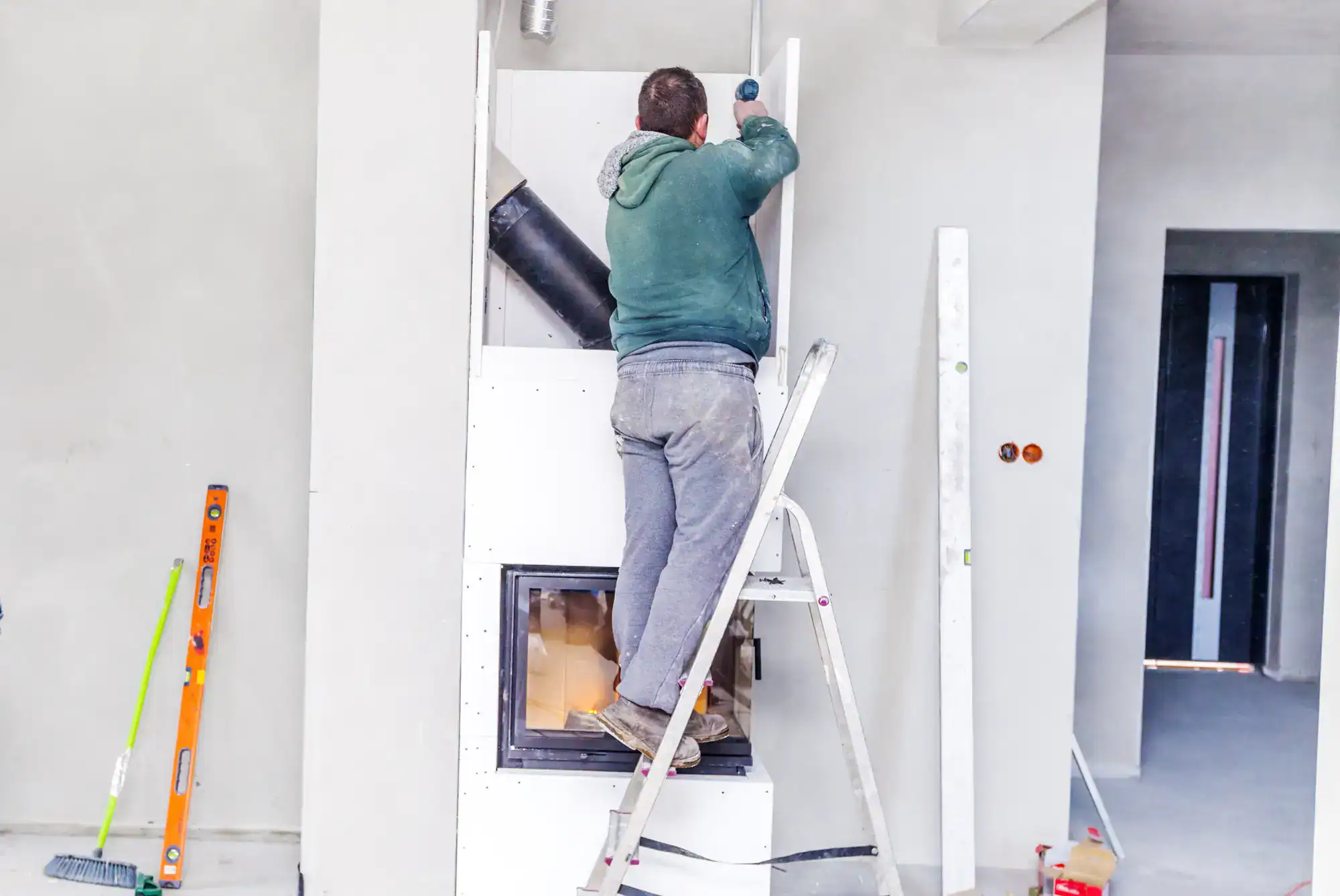 A person in a green hoodie stands on a stepladder, installing or fixing a white built-in fireplace in a room under construction—tools like a level and broom nearby—showcasing the skills of a masonry contractor Nassau County, NY.